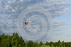 Colorful red and yellow hot air balloon flying over the green forest against a deep blue sky and clouds.