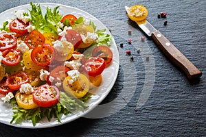 Colorful salad, fresh green leaves and sliced red and yellow cherry tomatoes, white plate, knife, black stone background