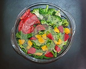A colorful salad, inside a round pyrex, with various vegetables and fruits such as tomatoes, broccoli, lettuce, arugula,