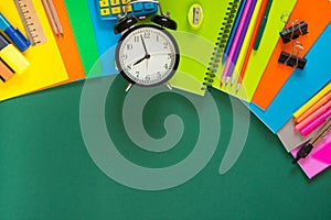 Colorful school supplies, book, and alarm clock on green. Top view, flat lay, copy space.