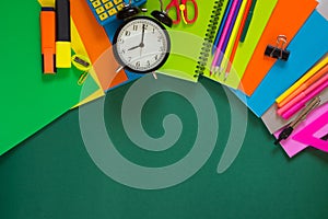 Colorful school supplies, book, and alarm clock on green. Top view, flat lay, copy space.