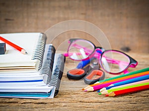 Colorful school supplies with books, color pencils, pink glasses, pen and cutter on wooden background.