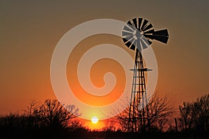 Colorful sky that`s orange and yellow with tree`s and a Windmill silhouette out in the country in Kansas.