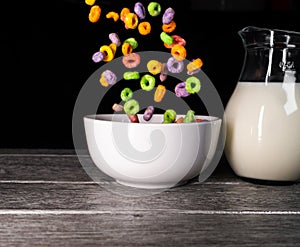 Colorful sugary cereals falling into a white bowl and milk jug in the background