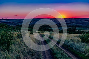 A colorful sunset over a clear sky of unusual color, around uneven large and spacious fields with green vegetation