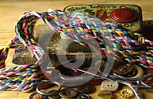 COLORFUL THREAD BRAID ON OLD METAL PIN BOX WITH BUTTONS AND SEWING AIDS IN THE BACKGROUND