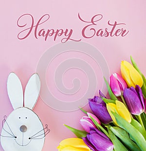 Colorful tulips on soft pink ground with white wooden easter bunny and happy easter text