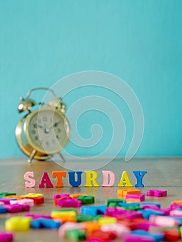 Colorful wooden word SATURDAY on wooden table and vintage alarm clock and background is powder blue. English alphabet made of wood