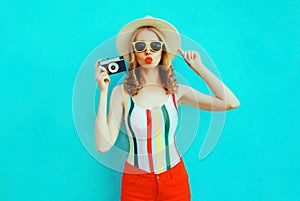 Colorful young woman holding retro camera, blowing red lips sends air kiss in summer straw hat having fun on blue wall