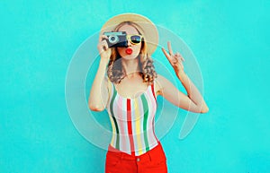 Colorful young woman holding retro camera, blowing red lips sends air kiss in summer straw hat having fun on blue wall