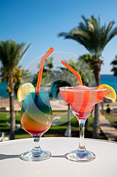 Colourful cold Rainbow Paradise and Strawberry daiquiri cocktail drinks served in glasses at pool bar overlooking blue pool, sea