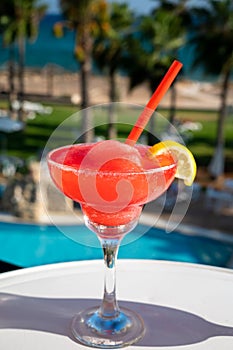 Colourful cold Strawberry daiquiri cocktail drink served in glass at outdoor cafe overlooking blue pool, sea and palm trees, relax