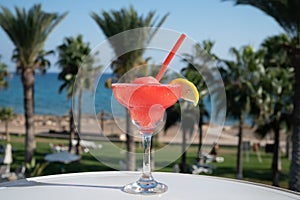 Colourful cold Strawberry daiquiri cocktail drink served in glass at outdoor cafe overlooking blue sea and palm trees, relax and