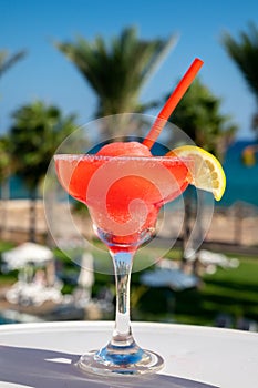 Colourful cold Strawberry daiquiri cocktail drink served in glass at outdoor cafe overlooking blue sea and palm trees, relax and