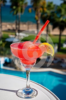 Colourful cold Strawberry daiquiri cocktail drink served in glass at pool bar overlooking blue pool, sea and palm trees, relax and