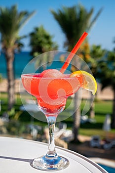 Colourful cold Strawberry daiquiri cocktail drink served in glass at pool bar overlooking blue pool, sea and palm trees, relax and