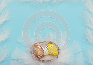 Colourful painted orange and yellow Easter eggs with Easter rabbit in basket with white feathers like a nest on blue background.