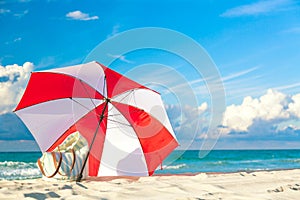 Colourful red and white umbrella with beach bag on the ocean beach against beautiful blue sky and clouds. Relaxation, vacation