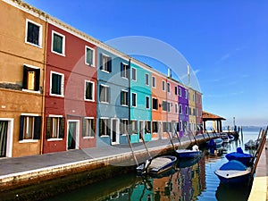 A colourful row of old homes along a canal and beside the sea, on a quiet sunny morning in the beautiful town of Burano, Italy.