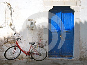 Colourful summer scene of an old red bike outside a greek house with whitewashed walls and a blue painted door in bright sunlight