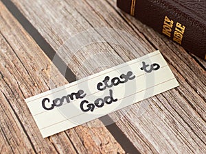 Come close to God Jesus Christ. A closeup of a handwritten quote and closed Holy Bible book on wooden background.