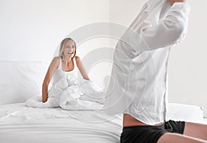 Come and join me. A young woman laughing while lying in bed and watching her boyfriend undress.