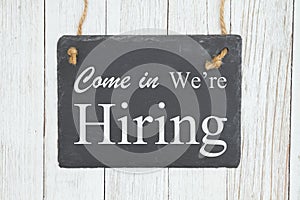 Come in we`re hiring text on a hanging chalkboard on weathered whitewash textured wood