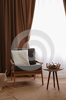 Comfortable armchair with cushion near window with stylish curtains in living room. Interior design
