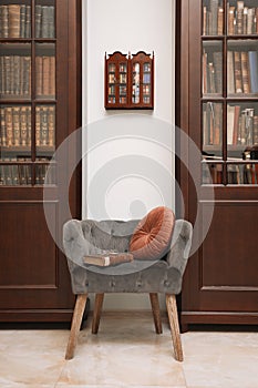 Comfortable armchair with pillow and book between wooden bookcases in library photo