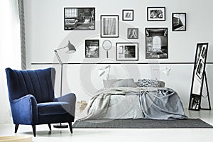 Comfortable blue armchair in monochromatic bedroom interior with king size bed and gallery of posters on the wall