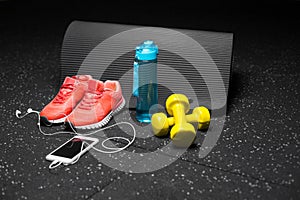 Comfortable sports shoes, a bottle of water, dumbbells, and phone on a black background. Accessories for gym training.