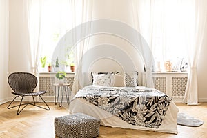Comfy bedroom interior with a leaf motif bedding on a bed, a rattan chair and a black and white pouf standing on a herringbone par
