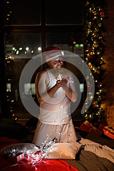 A comic image of santa claus in underwear on christmas eve. A man in a suit of Santa Claus holds a garland with lights