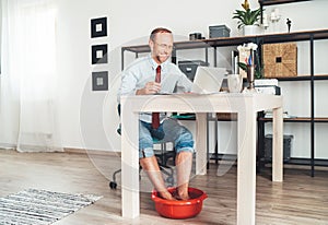 Comic modern office table situation. Businessman typing on laptop keyboard and soaring his feet in Foot hot Bath under table. Dis