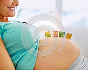 Coming soon. a pregnant woman lying down with wooden baby blocks on her belly at home.