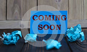 COMING SOON - words on blue paper on a dark wooden background with crumpled pieces of paper