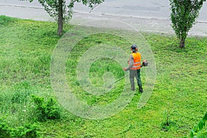 A communal service worker in an orange vest mows grass in a park with a hand gas mower