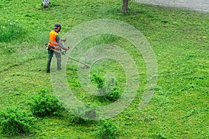 A communal service worker in an orange vest mows grass in a park with a hand gas mower