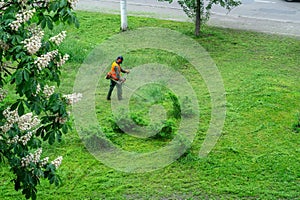 A communal service worker in an orange vest mows grass in a park with a hand gas mower