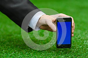 Communication and Business Subject: Hand in a black suit holding a modern phone with blue screen in the background of green grass