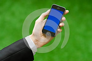 Communication and Business Subject: Hand in a black suit holding a modern phone with blue screen in the background of green grass