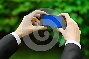 Communication and Business Subject: Hand in a black suit holding a modern phone with blue screen in the background of green grass