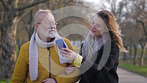 Communication of the granddad with the granddaughter during walk, happy girl with mobile phone shows photo to her old