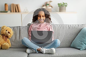 Communication and technology. Smiling black teen girl sitting on sofa and using laptop computer