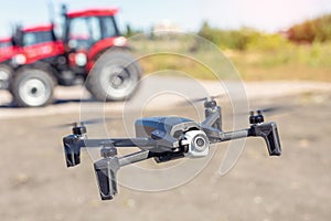 Compact black drone quadcopter with surveillance camera flying low against red agricultural tractor machines. Usage modern