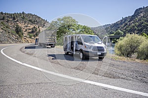 Compact mini van and big rig semi trucks with semi trailers take a break standing on the road shoulder on the bank of a mountain