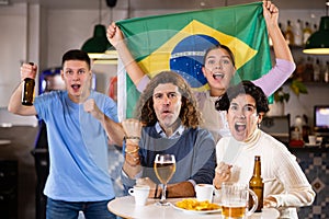 Company of emotional young adult fans supporting sports team of Brazil with state flag while resting with beer in bar