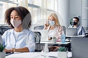 Company employees work in office. Multiracial managers in protective masks at workplaces with antiseptics and laptops