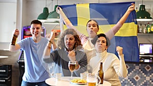 Company of excited young adult sports fans waving flag of Sweden and supporting national team with beer in bar