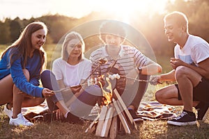 Company friends prepare roasted marshmallows snack isolated on nature background, youth group sitting around bonfire and